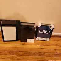 Photo of six black plastic photo frames arranged on a hardwood floor against a beige wall. Includes one sealed 8.5x11 inch frame with protective corner covers and other frames in various smaller sizes.