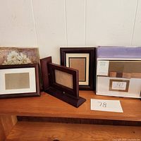Three empty wooden photo frames displayed on a wood surface against a wall. Two frames are dark-finished rectangular frames, and one is brown-finished with a stand. Included is a packaged photo book with 50 sleeves in front of the frames.