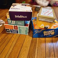 Photo of stacked boxed games showing littleBits Rule Your Room Kit, ThinkFun Smart Mouth, Dominoes in tin, and other card game boxes.