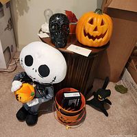 View of plush skeleton figure, black decorative skull, large ceramic pumpkin, small black cat figurine, and pumpkin bucket on carpet near wooden table and cardboard box