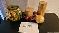 Photo showing four holiday decor items on a black surface: green glass candle holder decorated with raffia and gold leaves, two gold cylindrical candles, one standing and one on a red berry wreath, and a gold spherical candle.