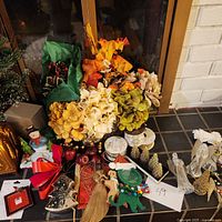 Overall assortment of holiday decorations including ornaments, figurines, artificial floral picks, and ribbons arranged on a tiled floor near a window and fireplace