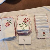Full set of white table linens laid out: 1 table runner, 6 napkins, 6 mini napkins, showing embroidered floral motifs and scalloped red edges.