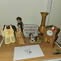 Full view of all small home decor items arranged on a glass shelf with a white paper labeled '46'. Features porcelain angel figurines, seated brown figurine, brass vase, clock, and others.