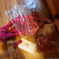 Photo showing multiple pink kids clothes hangers with metal hooks, some wrapped in plastic, a set of matching pink clips in a plastic bag, and a paper label with number 144.