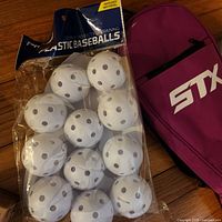 Pack of 12 white plastic practice baseballs with holes and pink STX lacrosse stick bag on wood floor.