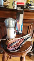 Photo showing Hamilton Beach hand blender, Sunbeam chopper, and various kitchen utensils on wooden stool with hand blender box in background