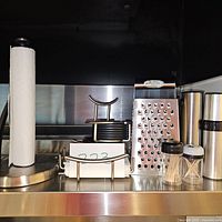 Photo showing paper towel holder, coaster stand with coasters, cheese grater, salt and pepper shakers, and thermos on kitchen countertop.