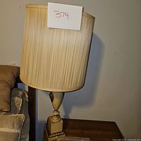 Vintage urn-shaped lamp with cream-colored ceramic base, ornate brass-colored handles and face decoration on a square pedestal base, with cream pleated fabric shade on top.