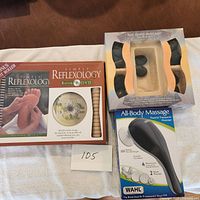 Photo showing Simply Reflexology kit box with book, DVD, and wooden foot roller alongside hot stone set and Wahl massager.