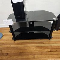 Full view of the black media cabinet showing three glass shelves with metal supports and open sides and back.