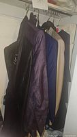Four men's suits hanging on hangers, showing fabric colors and textures including black, dark purple with pattern, navy blue with pattern, and beige.
