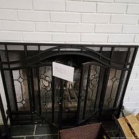 Front view of the metal two-door fireplace screen placed in front of a white brick fireplace. Shows the whole screen with glass floral panels and black metal mesh frame.