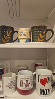 Three mugs on shelf including two with brown leaf patterns and one with 'Spill The Tea' text and colorful stripes