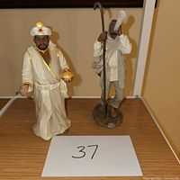 Two nativity figurines on wood surface: man with turban holding golden orb and man with staff and horn