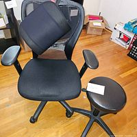 Photo of black mesh office chair with black lumbar back brace cushion attached and black padded armrests. Also shows a round black stool with caster legs next to the chair.