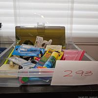 Clear plastic storage box showing assorted office supplies including glue sticks, pens, labels, and plastic folders inside.