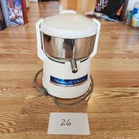 Photo of the full juicer showing white body and stainless steel container on wood floor with power cord underneath.