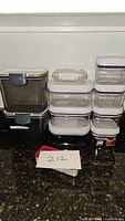 Photo showing 11 clear plastic storage containers, various sizes, with white lids and some with gray locking clips, stacked and arranged on a dark countertop.