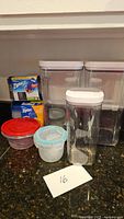 Photo showing three clear plastic storage containers with white lids, two closed small plastic containers, and two boxes of Ziploc bags.