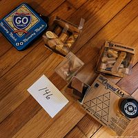 Photo showing 5 puzzle games on wooden floor including Memory Match tin, wooden block puzzles in plastic boxes, and The Original IQ Tester box with peg storage.
