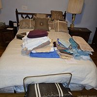 Full view of queen size bed with bedding piled on top, including decorative pillows and folded sheets and towels. Nightstands and lamps also partially visible.