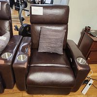 Brown leather electric reclining chair with armrest cup holders, a gray pillow, and visible controls for reclining on the right armrest.