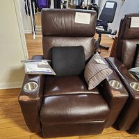 Front view of a brown leather electric reclining chair with armrests featuring built-in cup holders, two cushions placed on the seat, showing overall worn condition.