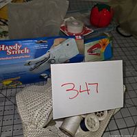 Photo shows various sewing items including Handy Stitch handheld sewing machine in box, spools of thread, buttons on cards, pin cushion shaped like tomato, and a knitted basket.