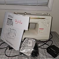 White Singer sewing machine on grid surface with instruction manual, foot pedal and power cord next to it, showing front side of machine and accessories.