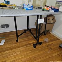 Full view of foldable sewing table showing black metal legs and caster wheels, tabletop surface includes various sewing supplies.