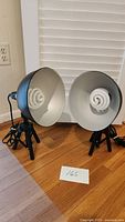 Two black Sharpics adjustable lights with tripods, each with a compact spiral bulb and silver reflector inside, placed on wooden floor near white blinds