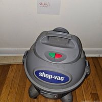 Gray and blue Shop-Vac wet/dry vacuum cleaner showing top handle and control switches, standing on wood floor against white wall.