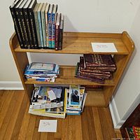 Small wooden bookshelf with three shelves filled with books and papers for scale. Bookshelf has a natural wood finish and shows in a corner of a room with hardwood floor.