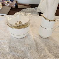 Photo showing white ceramic sugar bowl with matching lid, both with gold trim, positioned next to the creamer jug with gold trimmed handle and rim.