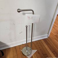Full view of brushed chrome toilet paper holder with vertical roll holder rod, standing on hardwood floor against white wall.