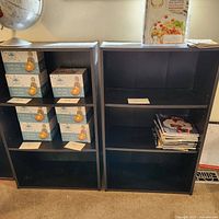 Two black laminate bookshelves standing side by side. One contains boxed items and the other has some magazines, but these are not included in the lot.