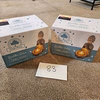 Two boxed OakiWay Buddha statue candle holders on carpeted floor with lot number card.