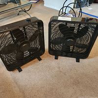Two black box fans positioned side by side on carpeted floor, showing front grille and blades.