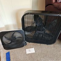 Two fans, one small desk fan and one larger box fan, both black plastic, shown on carpeted floor.