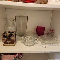 Six glass and decorative items on shelf: clear ribbed vase, tall square glass vase, clear cut glass bowl, clear handled dish, purple square vase, metal and bead candle holder