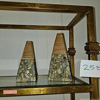 Photo of two tapered decorative vases with textured stone or concrete bases and grooved wooden-style tops on a glass shelf with gold metal frame.
