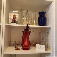 Wide photo showing various vases and figurines arranged on white shelving including blue and clear glass vases, a red vase, small figurines, and seashells.