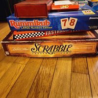 Stack of vintage board games including Scrabble, Rummikub Large Numbers Edition, Uno, Fast Track, and Dominoes in red case on top.