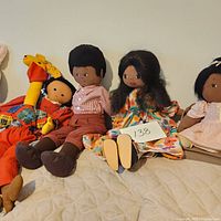 Full lot of five vintage stuffed dolls including three African-American cloth dolls, one giraffe doll, and one smaller doll in orange dress on a quilted surface.