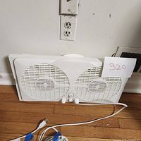 White window fan with dual blades and control knobs on a hardwood floor against a wall, showing front view and power cord coiled beside it.