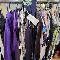 Photo of various women's tops hanging on a rack showing multiple colors and patterns including purple and floral prints.
