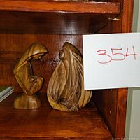 Two wooden hand-carved religious figurines displayed on a wooden shelf, one a praying woman and the other a veiled figure.