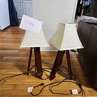 Pair of wooden tripod lamps on a hardwood floor with off-white rectangular shades and black power cords.