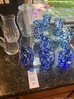 Full lot view showing 8 blue/white speckled glass vessels and 3 clear glass pitchers on counter.